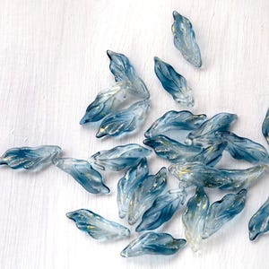 May include: A collection of translucent blue glass leaf-shaped beads with gold accents. The beads are scattered on a white surface, showcasing their delicate, organic forms. These beads are ideal for jewelry making and craft projects.
