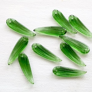 May include: Ten translucent green glass beads are displayed. Each bead is teardrop-shaped with vertical ridges and a small hole at the top. The beads are scattered on a white surface.
