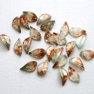 May include: A collection of small, marquise-shaped gemstones featuring a translucent appearance with swirling inclusions of copper, brown, and pale green hues. Each individual piece displays a smooth, polished surface with a curved, leaf-like profile.
