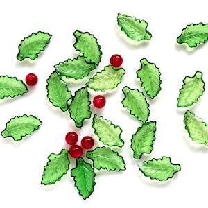 May include: Scattered on a white surface are translucent green leaf-shaped beads and small red round beads. The leaves have a detailed veining pattern. The beads are likely for crafting or jewelry making.