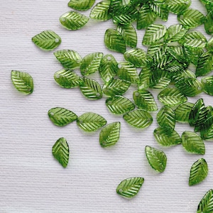 May include: A scattering of translucent green leaf-shaped beads on a white surface. Each bead features detailed veining and a small hole for threading. Suitable for jewellery making or craft projects.