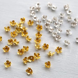 Könnte beinhalten: Eine Ansammlung kleiner, blütenförmiger Metallperlen in Gold- und Silbertönen. Die Perlen sind auf einer hellgrauen Oberfläche verstreut und zeigen ihre zarten Blütenblattdesigns. Ideal für Schmuckherstellung und Bastelarbeiten.