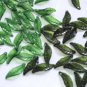 May include: A collection of translucent green leaf-shaped beads. The beads have detailed veining and a glossy finish, scattered across a white surface. The leaves vary in shade from light to dark green.