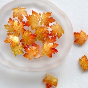 Könnte beinhalten: Eine Sammlung kleiner, durchscheinender Ahornblatt-Verzierungen in Gelb-, Orange- und Rottönen. Einige Blätter sind in einer Schale aus mattiertem Glas angeordnet, einige sind in der Nähe verstreut. Die Blätter haben eine glänzende Oberfläche.