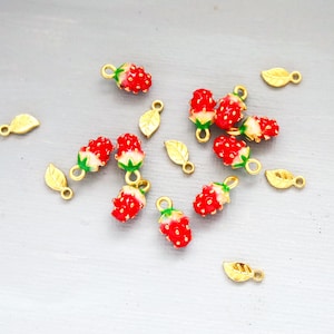 May include: A collection of small, red strawberry charms with green tops and white accents, scattered among gold-colored leaf charms. The strawberry charms have a loop for attaching to jewelry or other crafts.