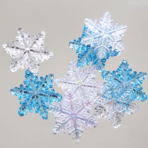 Op de afbeelding: Een verzameling heldere en blauwe sneeuwvlokornamenten. De ornamenten zijn gemaakt van een doorschijnend materiaal, met ingewikkelde details. De sneeuwvlokken variëren in grootte en zijn gerangschikt op een reflecterend oppervlak.