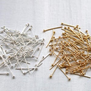 May include: A collection of silver and gold-colored head pins. The pins have a small ball at the top and a long, thin shaft. The pins are scattered on a light-colored surface, suitable for crafting and sewing projects.
