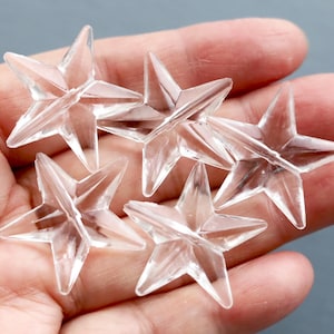 May include: Five clear, star-shaped decorative elements. Each star has five points and a faceted design, creating a sparkling effect. The stars are held in a hand against a gray background. These could be used for crafts or decorations.