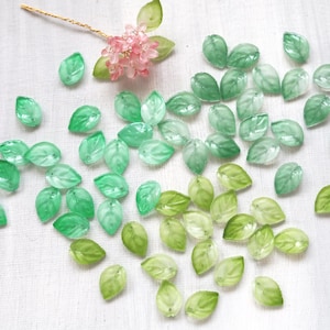 May include: A scattering of green leaf-shaped beads on a white background. The beads are in various shades of green. A small floral element with pink petals and a gold stem is also visible, suggesting a nature-inspired theme for crafting or jewellery.