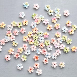 Peut inclure: Une collection de petites décorations en forme de fleurs aux couleurs pastel, dispersées sur une surface grise. Les fleurs présentent une variété de couleurs, notamment rose, jaune, bleu et vert, avec un centre plus foncé. Idéal pour les projets d'artisanat.
