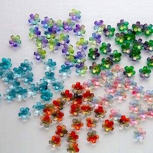 Könnte beinhalten: Eine Ansammlung durchscheinender, blütenförmiger Perlen in verschiedenen Farben, darunter Blau, Grün, Lila, Rot und Orange. Die Perlen sind auf einer weißen Oberfläche verstreut und bilden eine farbenfrohe und dekorative Darstellung. Ideal für Bastelarbeiten und Schmuckherstellung.
