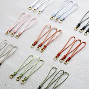 May include: A collection of colourful wrist straps with gold-coloured metal rings. The straps are in various colours, including pink, brown, green, and black. Each strap has a looped design and a woven detail near the ring.