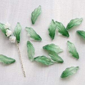 May include: A collection of green glass leaves and a stem with white flower buds. The leaves are translucent and vary in size. The stem is silver-colored and twisted. The white flowers have a pearl-like center.