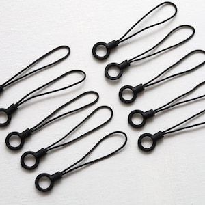 May include: A collection of black phone charms with a loop and ring design. The charms are made of a flexible material and are arranged on a white surface. These accessories are designed to attach to a phone or other small device.