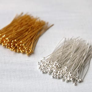 May include: Two bundles of head pins, one gold and one silver. Each pin features a small, round bead at the head and a long, thin wire stem. The pins are displayed on a white surface, suitable for craft projects.