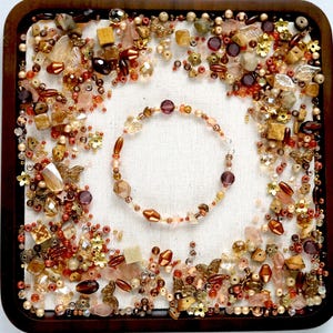 Peut inclure: Un plateau carré en bois rempli d'un assortiment de perles, de pierres précieuses et d'éléments décoratifs dans les tons or, marron et rose. Un bracelet de perles avec des perles violettes et dorées est au centre. Le plateau est sur une surface blanche.
