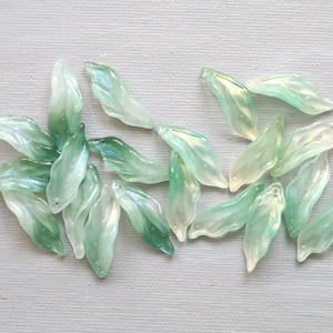 May include: A selection of translucent, leaf-shaped beads in shades of green and white. The beads have a subtle iridescent sheen, scattered on a light grey surface. These beads are suitable for jewellery making and craft projects.