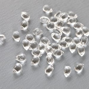 May include: A collection of clear, teardrop-shaped beads scattered on a gray surface. The beads are translucent, reflecting light, and appear to be made of glass or a similar material. The image is a close-up, highlighting the texture and shape of the beads.