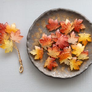 Peut inclure: Des décorations de feuilles d'érable colorées. Certaines sont disposées sur une assiette grise foncée avec des accents dorés, d'autres sont fixées à une tige en fil d'or. Les feuilles présentent des nuances de rouge, d'orange et de jaune.