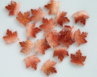 May include: A collection of small, maple leaf-shaped embellishments. The leaves are a gradient of orange and red with a glittery finish. These decorative elements are ideal for crafting and seasonal projects.