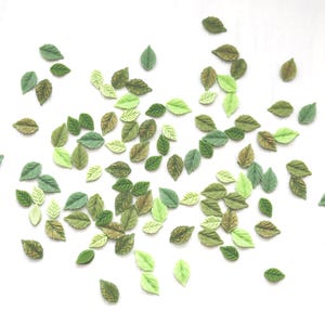 May include: A scattering of miniature artificial leaves in shades of green and brown against a white backdrop. The leaves range from pale to deep green, with some showing textured patterns, creating a naturalistic effect.
