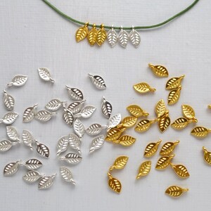 May include: A collection of gold and silver leaf-shaped charms. Some are strung on a green cord, others are loose. These charms are suitable for jewellery making or craft projects.