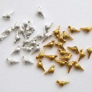 Op de afbeelding: Een verzameling kleine, vogelvormige bedels in zilver en goud op een witte achtergrond. De zilveren bedels zijn gegroepeerd, terwijl de gouden bedels apart liggen. Deze bedels zijn ideaal voor het maken van sieraden en andere knutselprojecten.