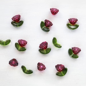 May include: A collection of small, decorative glass beads in the shape of flowers. Each flower features a deep red, faceted bead for the bloom, set atop green, leaf-shaped beads. The beads are scattered on a white surface.
