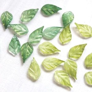 May include: A collection of small, translucent glass leaves in various shades of green and yellow-green. The leaves have detailed veining and are scattered on a white surface, suggesting a craft or decorative item.