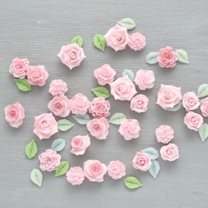 May include: A collection of small, light pink rose blossoms and green leaves are scattered on a gray surface. The roses vary in size and are arranged in a visually appealing pattern, suggesting a craft or decorative theme.