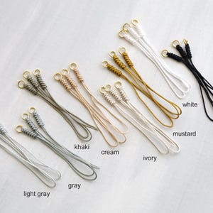 May include: A collection of handmade jewelry tassels in various colors, including light gray, gray, khaki, cream, ivory, mustard, white, and black. Each tassel features a gold-colored ring at the top and is made of braided cord.