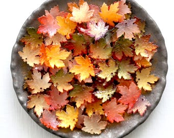 May include: A collection of colorful, translucent maple leaf-shaped beads in shades of red, orange, yellow, and green. The beads are arranged on a dark gray, textured plate, suggesting a fall theme.