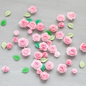 May include: A collection of small, pink rose flower embellishments and green leaf accents scattered on a light gray surface. The roses vary in size and are in full bloom, with detailed petals. The leaves are a vibrant green.