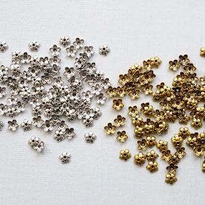 May include: Two piles of small, flower-shaped metal bead caps, one silver, one gold. The bead caps are scattered on a light surface, showing the contrast between the metallic finishes and the background.