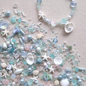 May include: A beaded bracelet featuring white starfish, light blue and pink translucent beads, and small seashell charms, surrounded by a scattered collection of loose craft beads in shades of aqua, pearl, white, and soft pink.