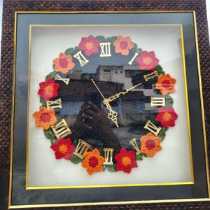 May include: A framed wall clock with a dark brown woven frame. The clock face features a crochet floral design in red, orange, and green, surrounding gold Roman numerals. The clock hands are gold, and the background shows a blurred image.