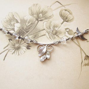 May include: A silver-toned necklace featuring a delicate bird charm and a leaf pendant with clear crystal accents. The necklace has a chain with clear beads and is set against a vintage floral background.