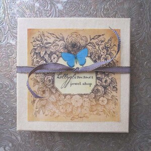 May include: A small, white box with a brown ribbon and a blue butterfly design. The box has a label that says "Hollyglimmer Jewel Shop".