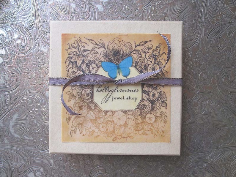 May include: A small, white box with a brown ribbon and a blue butterfly design. The box has a label that says "Hollyglimmer Jewel Shop".