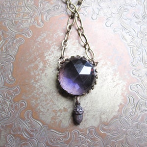 May include: A purple glass pendant necklace with an acorn charm hangs from a brass chain. The necklace is displayed on a patterned metal surface.