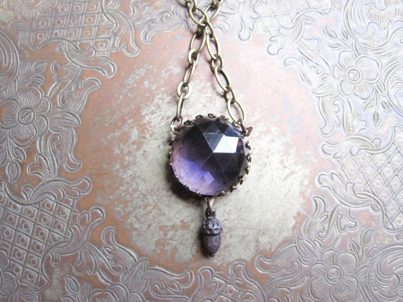 May include: A purple glass pendant necklace with an acorn charm hangs from a brass chain. The necklace is displayed on a patterned metal surface.