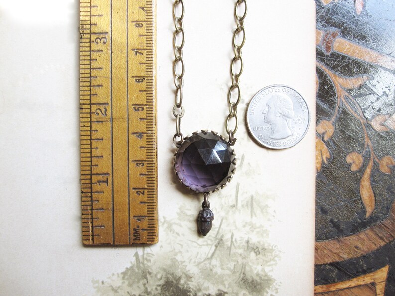 May include: A purple glass pendant necklace with a brass chain and an acorn charm. The pendant is faceted and has a geometric design. The necklace is approximately 18 inches long.
