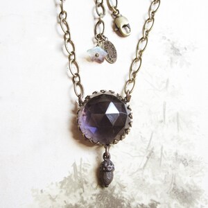 May include: A purple faceted glass pendant necklace with a brass chain and an acorn charm. The pendant is set in a brass frame and has a star charm attached to the chain. The tag on the chain reads "almost there".