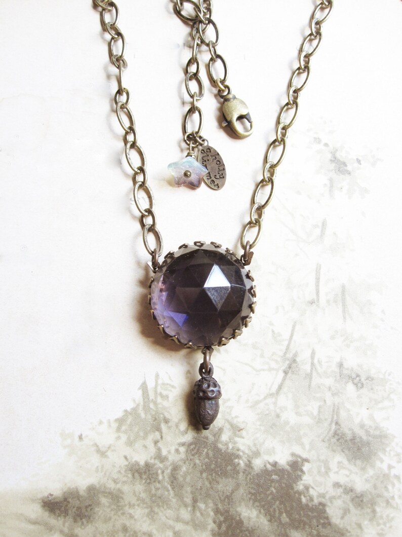 May include: A purple faceted glass pendant necklace with a brass chain and an acorn charm. The pendant is set in a brass frame and has a star charm attached to the chain. The tag on the chain reads "almost there".