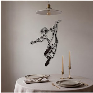 Könnte beinhalten: Ein Wandkunstobjekt aus schwarzem Metall, das eine stilisierte Figur in einer dynamischen Sprungpose mit ausgestreckten Armen zeigt. Die Figur trägt weite Kleidung und ist an einer schlichten Wand über einem runden Esstisch montiert.