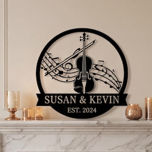 May include: Black metal wall art featuring a violin, bow, and musical notes within a circular frame. The names "SUSAN & KEVIN" are displayed on a banner, with "EST. 2024" below. The artwork is set against a neutral wall.