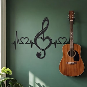 Può includere: Chitarra acustica appesa a una parete verde accanto a un'opera d'arte murale in metallo nero. L'opera d'arte presenta una chiave di violino a forma di cuore, collegata a una linea di battito cardiaco con altri due cuori.