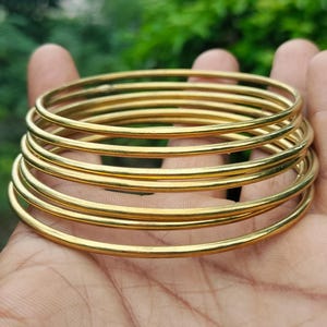 Peut inclure: Une pile de bracelets de couleur or, chacun avec une surface lisse et arrondie. Les bracelets sont tenus dans une main, avec un arrière-plan vert flou. Les bracelets semblent être en métal et ont une finition brillante.