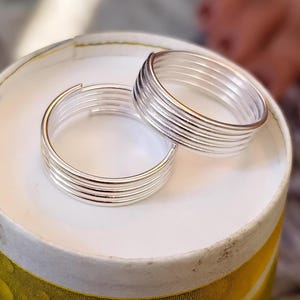 May include: Two silver-colored rings, each constructed from a single continuous wire coiled into five parallel loops. The rings feature a smooth, polished metallic finish and an open-ended design, showcasing a layered, multi-band appearance.