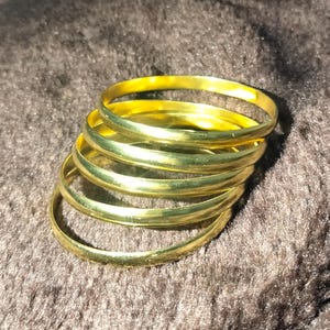Puede incluir: Una pila de seis pulseras de color dorado. Las pulseras son lisas y brillantes, reflejando la luz. Están apiladas unas sobre otras, creando un efecto de capas. El fondo es una superficie oscura y texturizada.